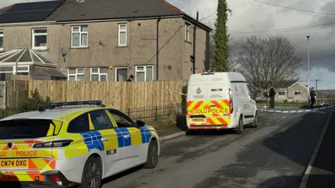 BBC A police car and police van are parked next to the police cordon on a road near to houses, with two police officers behind the cordon. 