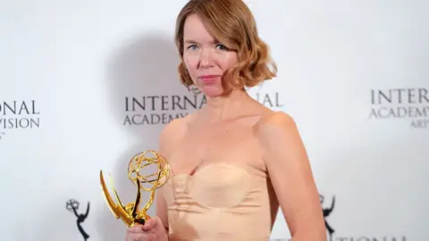 Getty Images Anna Maxwell-Martin stands in front of a backdrop covered in International Emmy branding. She is holding a large, shining gold trophy and is wearing a pale peach-coloured dress. She is looking directly at the camera, with a slight smile on her face.