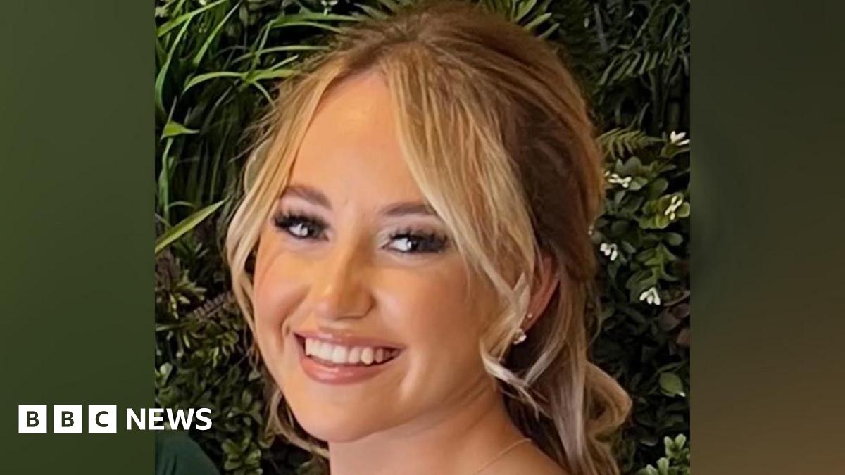 A woman, with blonde hair wearing makeup and fake eyelashes. She is smiling and standing in front of some green leaves.