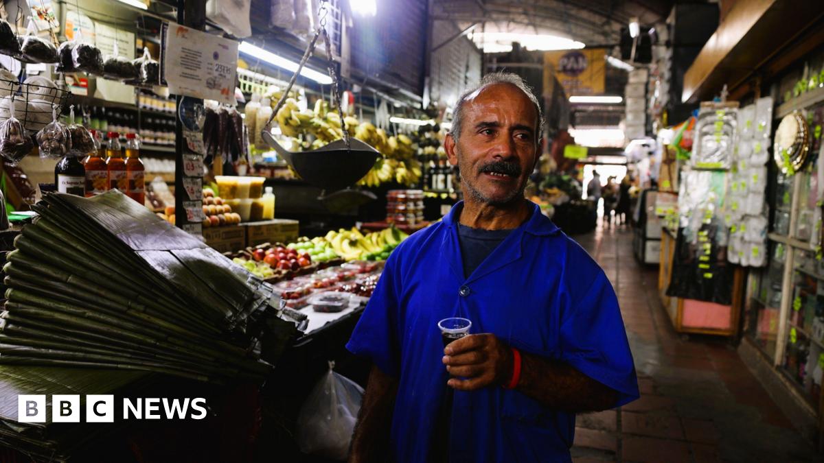 A man in a Caracas market speaks while holding a glass of coffee