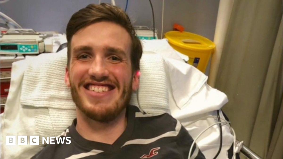 A young man with short dark hair and stubble looks directly at the camera in front of a dark blue background.
