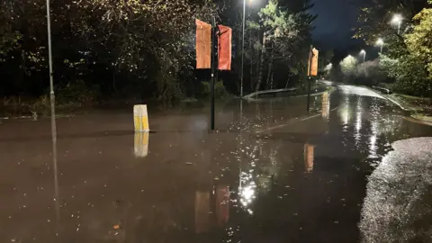 Water flooding the road on Piper's Way, which is empty of cars. It is dark and the streetlights are reflecting on the water. 