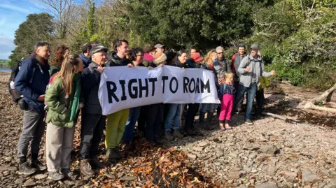 BBC A group of people stood together holding a banner which says RIGHT TO ROAM. They are standing on the banks of a river and are wearing backpacks and coats.
