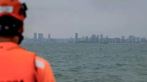 Getty Images A Taiwanese coast guard in orange attire looks out to sea. In the distance is the a city skyline.