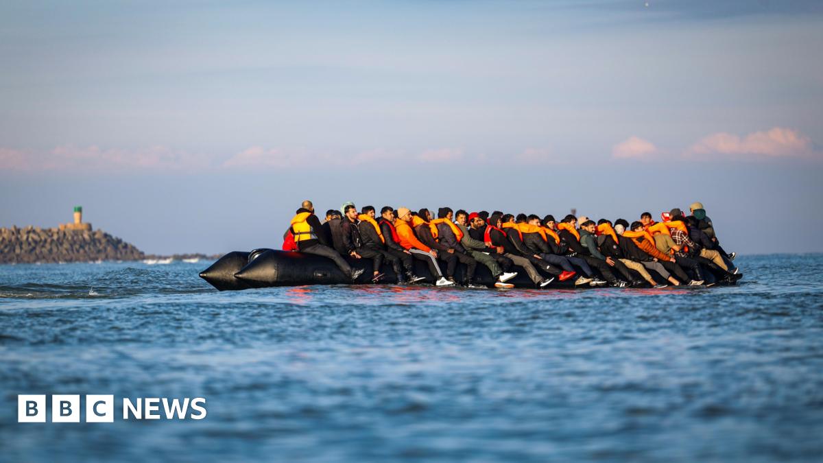 Migrants wearing orange lifevests sit on an inflatable boat on the English Channel, a rocky headland appears in the backdrop