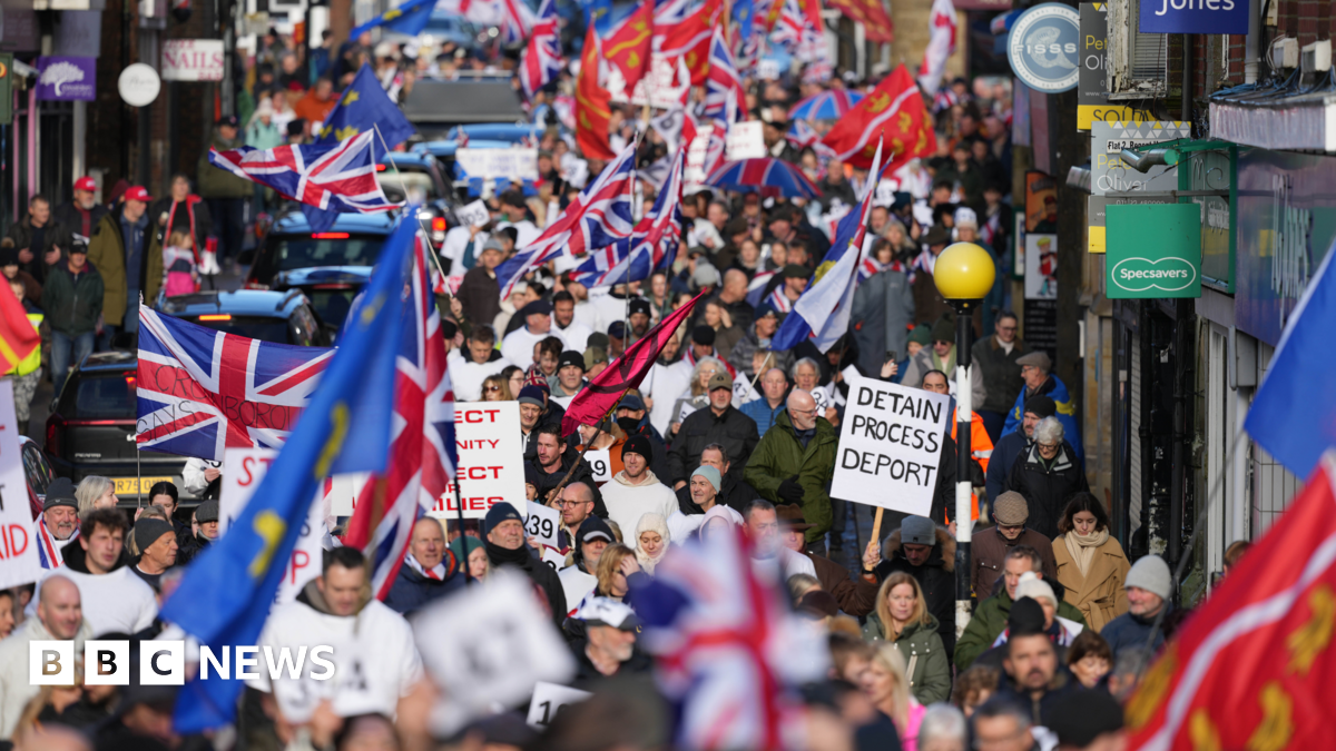 Hundreds of people marching on a town high street. Some people are carrying union jacks and Sussex county flags, while one person holds a sign that reads "detain process deport".