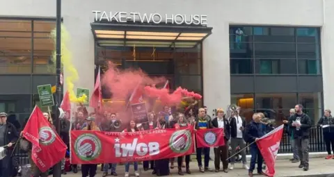 IWGB A crowd of people outside an office holding red and white banners which say IWGB and using red flares.