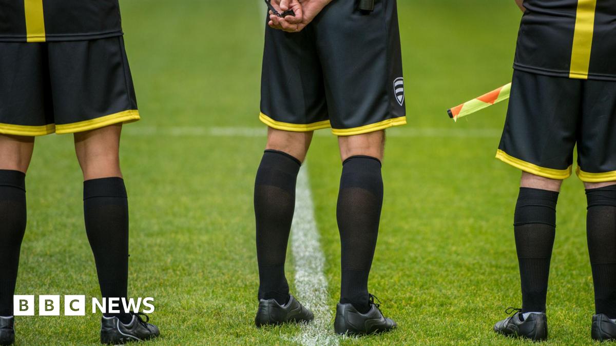The lower bodies of three football match officials standing on grass. The photo is taken from behind and they are wearing black and yellow kit.