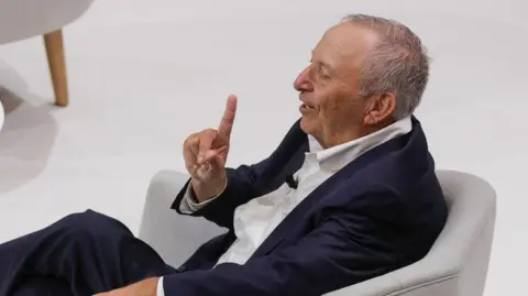 EPA Larry Summers, sat in profile on a grey armchair, wearing a navy blue suit and white short, smiles as he raises the index finger of his right hand