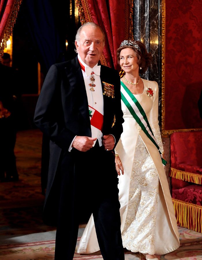 Former King Juan Carlos in formal attire with medals, walking beside Queen Letizia in a white gown with a green sash.