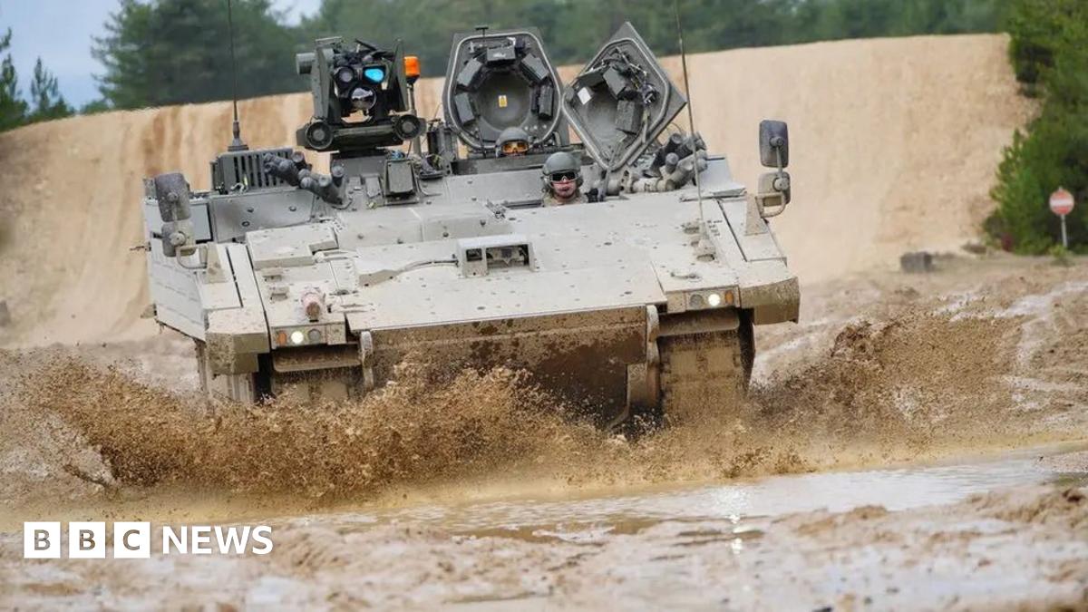 An Ajax armoured vehicle being driven through a muddy terrain. Mud is being kicked up off the surface in all directions around the vehicle. Soldiers can be seen controlling the vehicle.