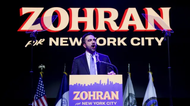 Zohran Mamdani in a dark suit stands behind a podium bearing the sign "Zohran for New York City" as he speaks at an election night party. Behind him are several flags, including the US flag to his right