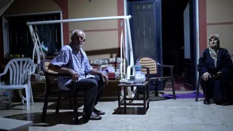 Neha Sharma/BBC An older man and woman sit on a tiled area outside a house, after dark.