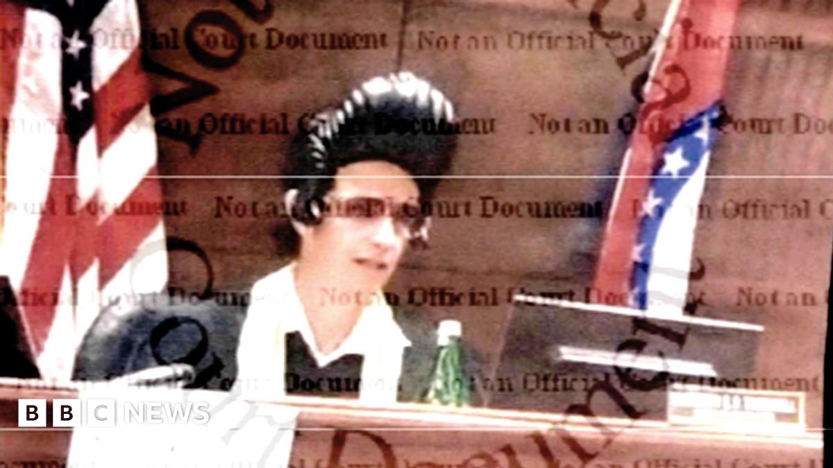 An image of Judge Thornhill sitting at the court bench, taken from official court documents, showing him wearing an Elvis wig with the singer's signature black pompadour and sunglasses, as well as a traditional judge's robe with a white scarf around his neck. In front of him is a computer screen and a bottle of water. Behind him is a flag of the US and a flag of the State of Missouri.