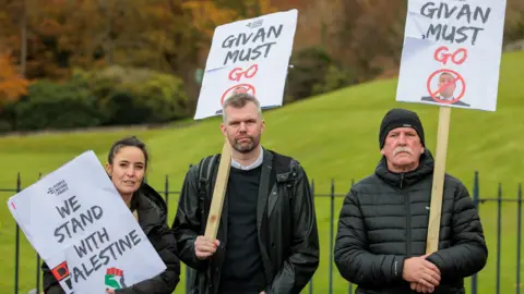 Liam McBurney/PA Wire Gerry Carroll stands with two protesters, a man and a woman, during a demonstration at Stormont.  They are all wearing dark-coloured coats and carrying placards.  Carroll, who has short, greying hair and beard, is carrying a "Givan Must Go" placard, as is the man beside him who has a grey moustache and a black beanie hat.  The woman, who has dark hair tied back, is holding a "We Stand With Palestine" placard.  There are trees and a green lawn in the background.