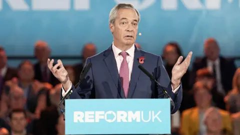 PA Media Nigel Farage in a suit and tie standing behind a podium with the words ‘REFORM UK’ printed on the front, speaking into two microphones. The background shows a large blue banner with partial text and an audience seated behind.