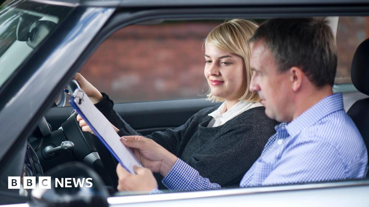 A young woman taking a driving test. She has blonde hair and is wearing a dark grey jumper. Her tester is a man in a check blue shirt. He is showing her a document on a clipboard.