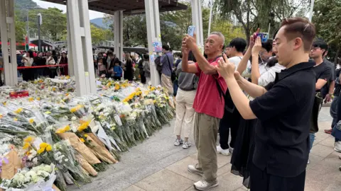 Ching Sze Yip/BBC Hundreds of flower bouquets on the left. On the right are people who are queuing, some of whom are taking photos