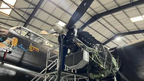 BBC/Karl Bird A side view of Just Jane in a warehouse. The aircraft's engine is exposed, revealing mechanical parts, pipes and wiring. 