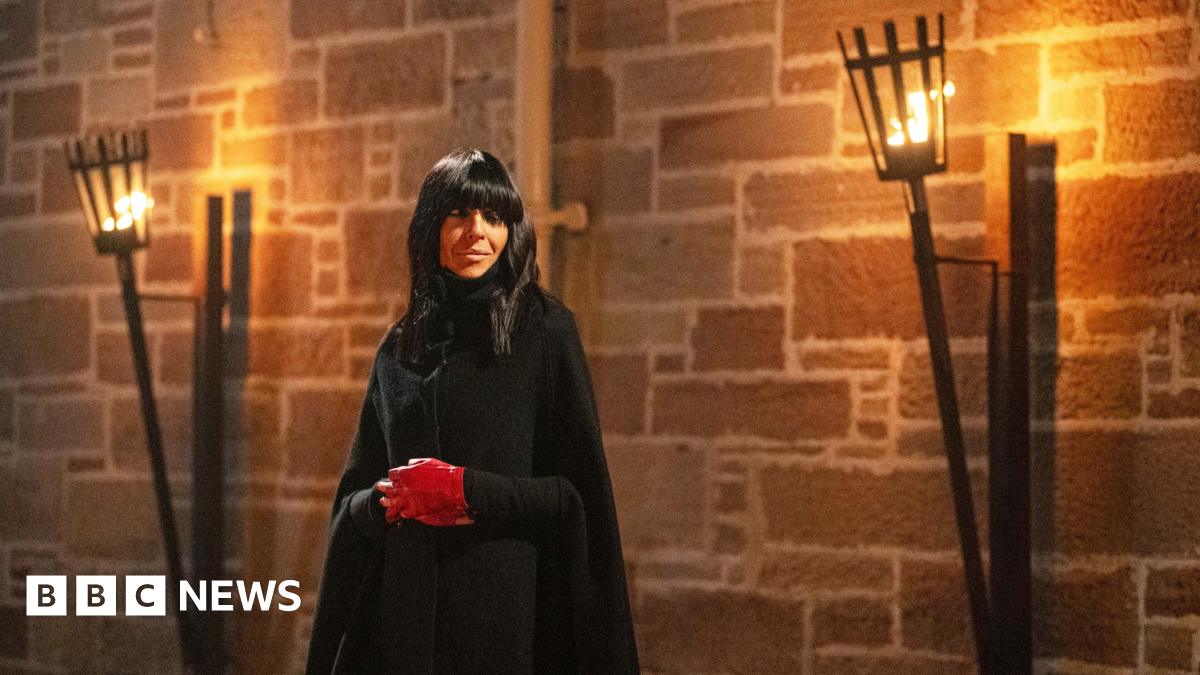 Picture of Claudia Winkleman in The Celebrity Traitors. She is in a black cloak and wearing red gloves. There arelanterns with flames behind her