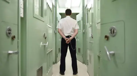 PA Media An officer stands with his back to the camera and his hands clasped behind him inside a prison. On either side there are light green cell doors.