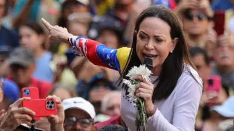 Getty Images Maria Corina Machado, a lady with brown hair and wearing a white t-shirt holds a microphone with flowers as a crowd stands behind her.