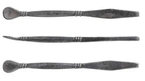 Norfolk County Council A silver stick with a spoon shape on one end and a rectangular end the other. Three round circles wrap around each end. The object is photographhed three times, the middle image shows it from a side angle.