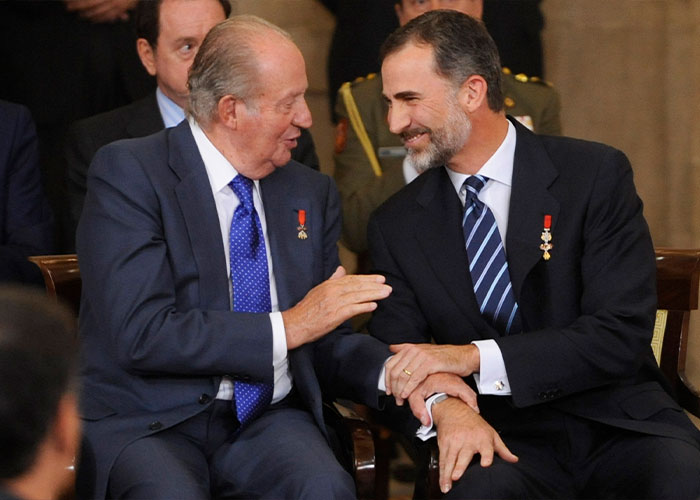 Former King Juan Carlos in a navy suit speaking to a man in a black suit during a formal event indoors