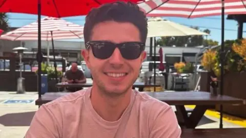 Mason Leposavic Mason Leposavic smiled while wearing sunglasses, sitting in a spot with picnic tables and umbrellas