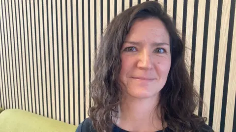 Mousumi Bakshi/BBC Jessica Rundell who is sitting on a light greeny yellow sofa against a wooden panelled wall. She has dark brown gently curling hair falling to her shoulders and is wearing a dark T-shirt. 