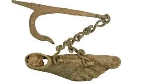 Kent County Council A rusted copper alloy carving of a foot, toenails included, attached to a diagonal chain hanging from a slim bar with a curved fork at the end