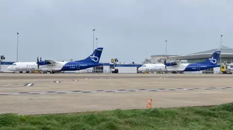 Two Blue Islands planes parked up on the tarmac at an airport. The terminal building is in the background of the shot.