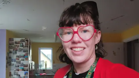 Kerrie poses for a selfie in the kitchen of her home, wearing a red cardigan and black printed blouse, she has a broad smile and striking heart-shaped spectacles