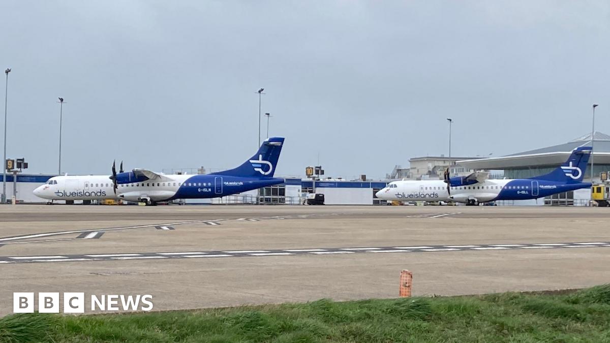 A shoulder shot photo of a man wearing a navy top wearing glasses standing in front of a pink and light brown government building.