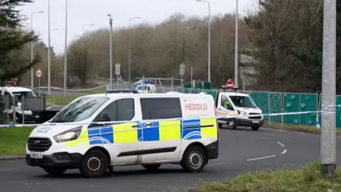 a police van is parked on a road with police tape and a barrier seen in the background.