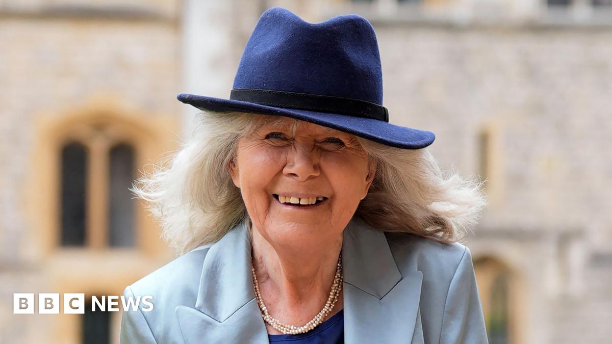 Jilly Cooper smiles at the camera. She has shoulder length grey hair and is wearing a light blue jacket and a dark blue top underneath, and a pearl necklace. She is also wearing a dark blue trilby-style hat