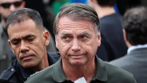Reuters/Adriano Machado Former Brazilian President Jair Bolsonaro wearing green polo shirt with police officer standing behind him