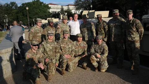 Getty Images A row of National Guardsmen in camouflage uniform kneel in front of a row of standing guards, with Dan Driscoll in white button-down shirt in the middle with his arms around two of them