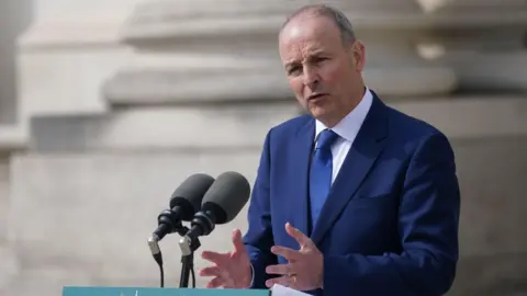 PA Media Micheál Martin is dressed in a blue suit, with a white shirt and navy tie. He is standing at a lectern. Two microphones are in front of him 