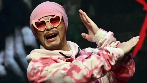 Getty Images Malaysian rapper Wee Meng Chee, known by his stage name Namewee, gestures during a press conference in Taipei. He is wearing a pink beanie, pink heart-shaped sunglasses, and a pink hoodie