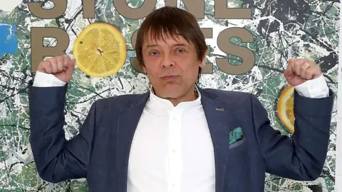 Getty Images Mani with both arms raise standing in front of a large image of the Stone Roses' debut album artwork