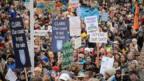 Getty Images Protestors demonstrating against the proposed XL-Northern Gateway pipeline project, at the British Columbia Legislature building in Victoria. The pipeline would bring crude oil from the Alberta tar sands through British Columbia to the pacific coast. The image shows hundreds of people holding signs that say "Defend our Coast" "Our Coast is not for Sale" and "You can't put a price on nature"