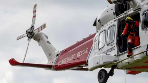 Getty Images HM Coastguard helicopter crew conduct a search and rescue training exercise over the cliff tops in England. It is a red and white helicopter with signage reading 'COASTGUARD RESCUE' on it.