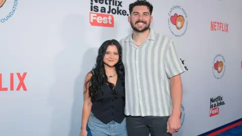 Getty Images for Netflix Kayla Vesia, wearing a black blazer and jeans, stands smiling next to husband Alex Vesia, wearing a button-down and jeans, in front of a Netflix step-and-repeat