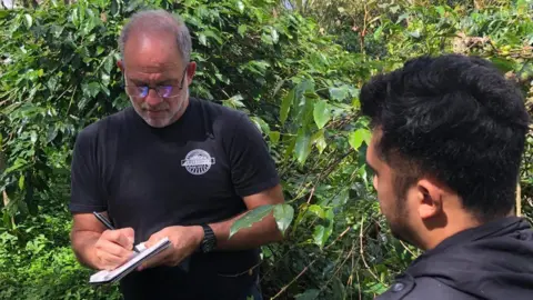 Cafe Campesino Pomeroy is wearing a black t-shirt and writing in a notebook with a black pen among green foliage, with the back of the head of a farmer in the foreground