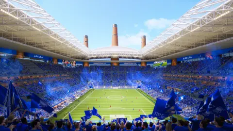 Birmingham City Inside the ground is a sea of blue where fans are seated in this artist impression. The pitch can be seen with players on it. Advertising screen surround the top ring around the ground which is partially covered with a white roof.