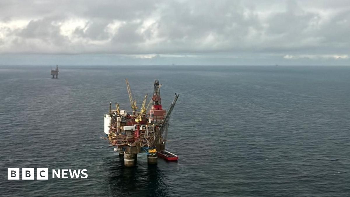 Two oil rigs, one in the foreground, and one in the distance, are seen sitting in a calm North Sea on a light but cloudy day.