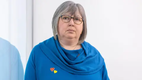 PA Media Brenda Doherty looking at the camera, she has grey hair and glasses and is wearing a blue dress with a heart pin. 