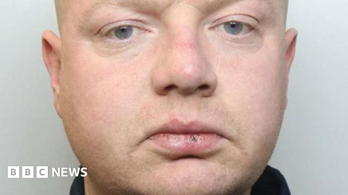 A close-up of a man's face. He has cracked lips, and is looking directly at the camera. He is wearing a dark top.