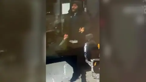 A man stands in a barbers' shop dressed in black with the hood of his coat over his head. He is gesticulating with one hand and the other one has a knife in it. He has a blue sweater on. 